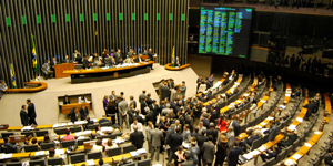 Fotografia colorida do Plenário da Câmara dos Deputados, em plano aberto, que mostra o Plenário lotado de pessoas, inclusive em volta da Mesa Diretora. À direita da imagem, um painel eletrônico de votação aparece em funcionamento.
